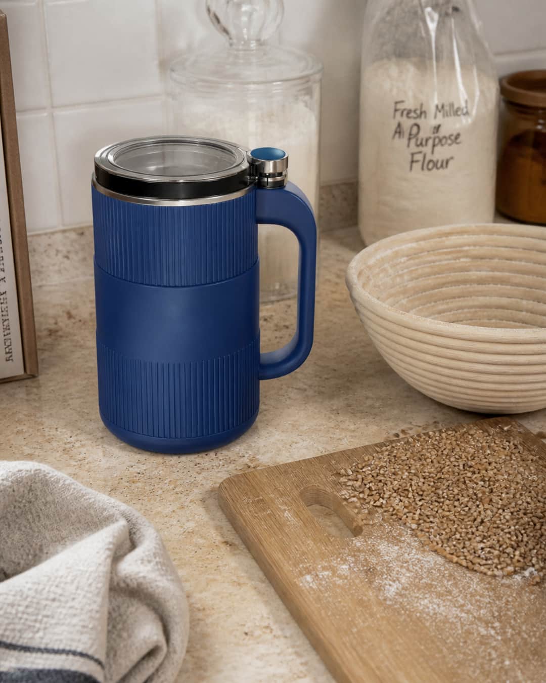 GRAIN BLOOM mill closed on a baking counter beside whole grain berries and a banneton