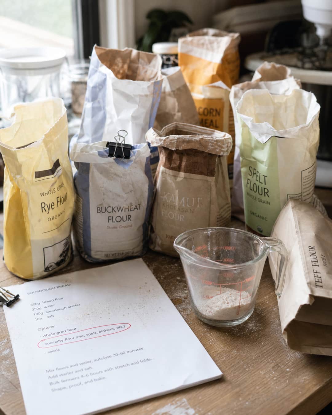 Home baker pantry with opened specialty flour bags, a recipe printout, and a measuring cup with only a little flour used