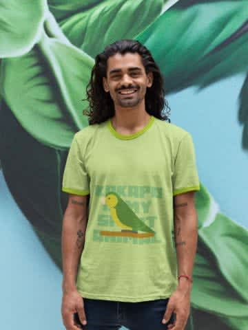 A brunette man with long brown hair, wearing a light green shirt with a parrot illustration while standing up against a green tropical leaves wallpaper 