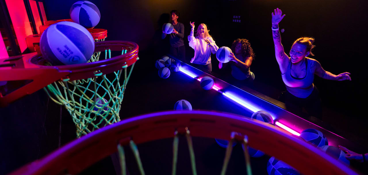 Four friends laugh and cheer while shooting glowing basketballs in Activate's neon-lit Hoops room.