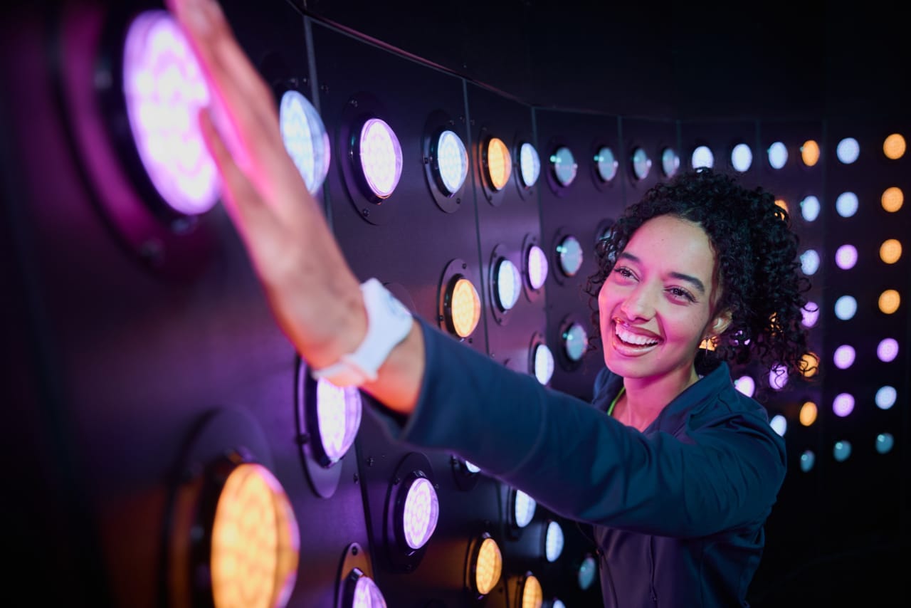 A player reaches up to press large glowing discs on the Press wall.