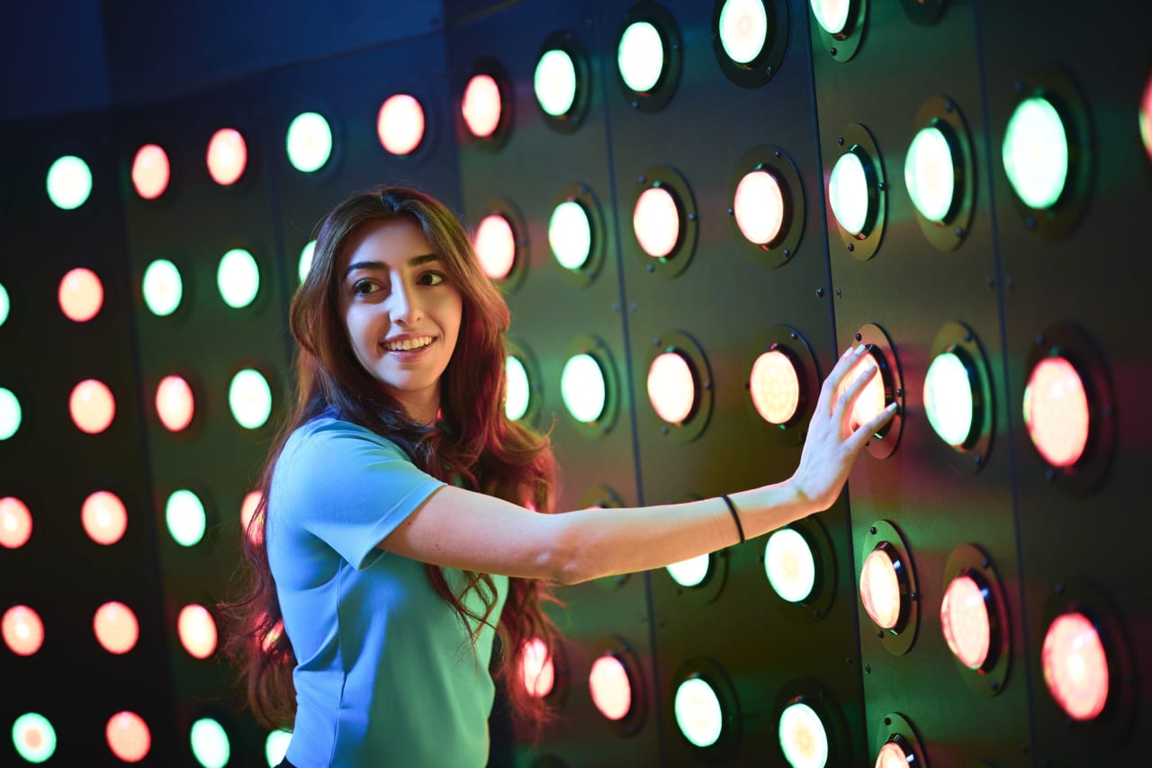 A player reaches for glowing buttons on a wall of coloured lights.