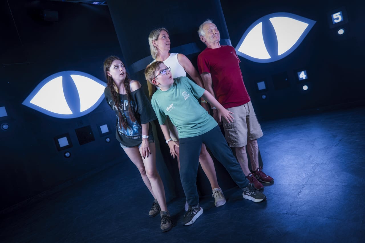 A family huddles together beneath large glowing eye graphics in the Hide room.