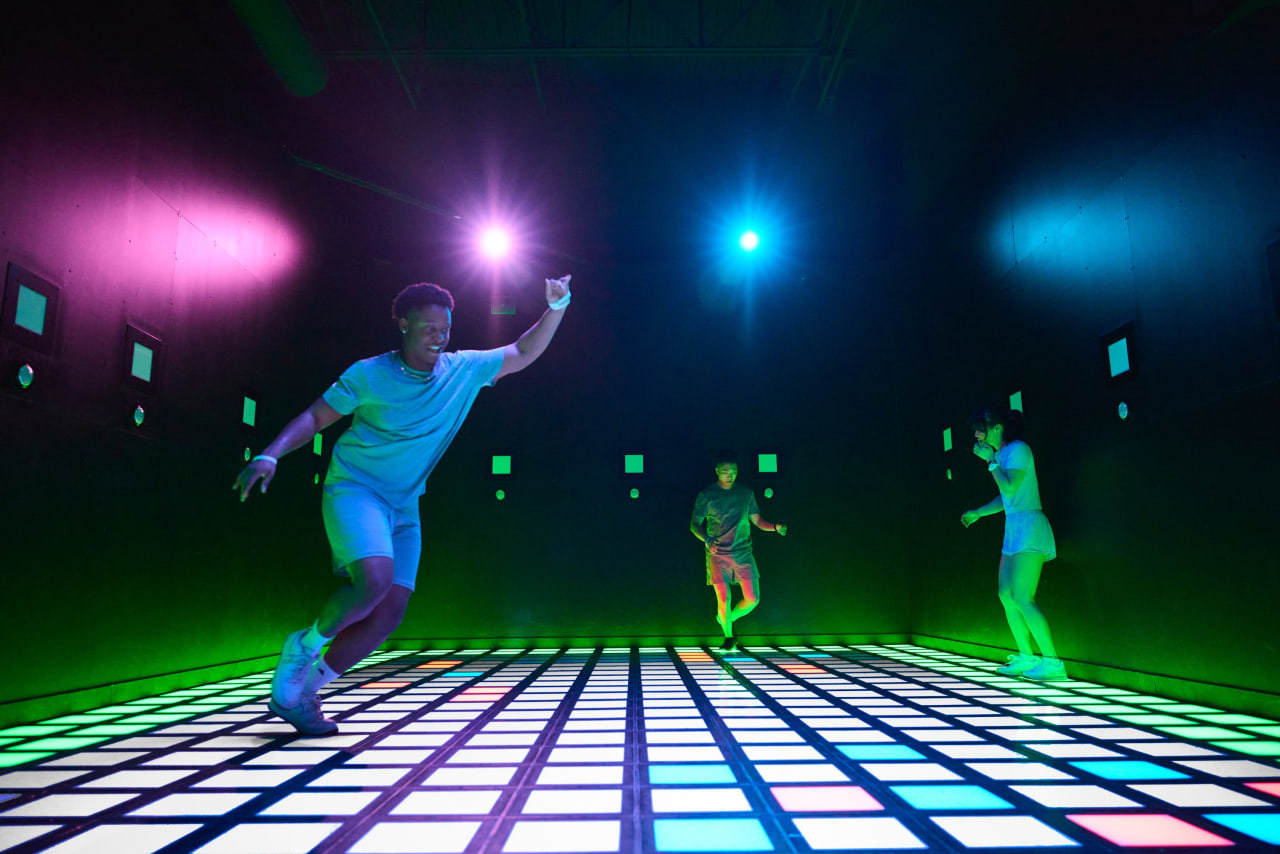 Players run across the Mega Grid's illuminated floor beneath coloured lights.