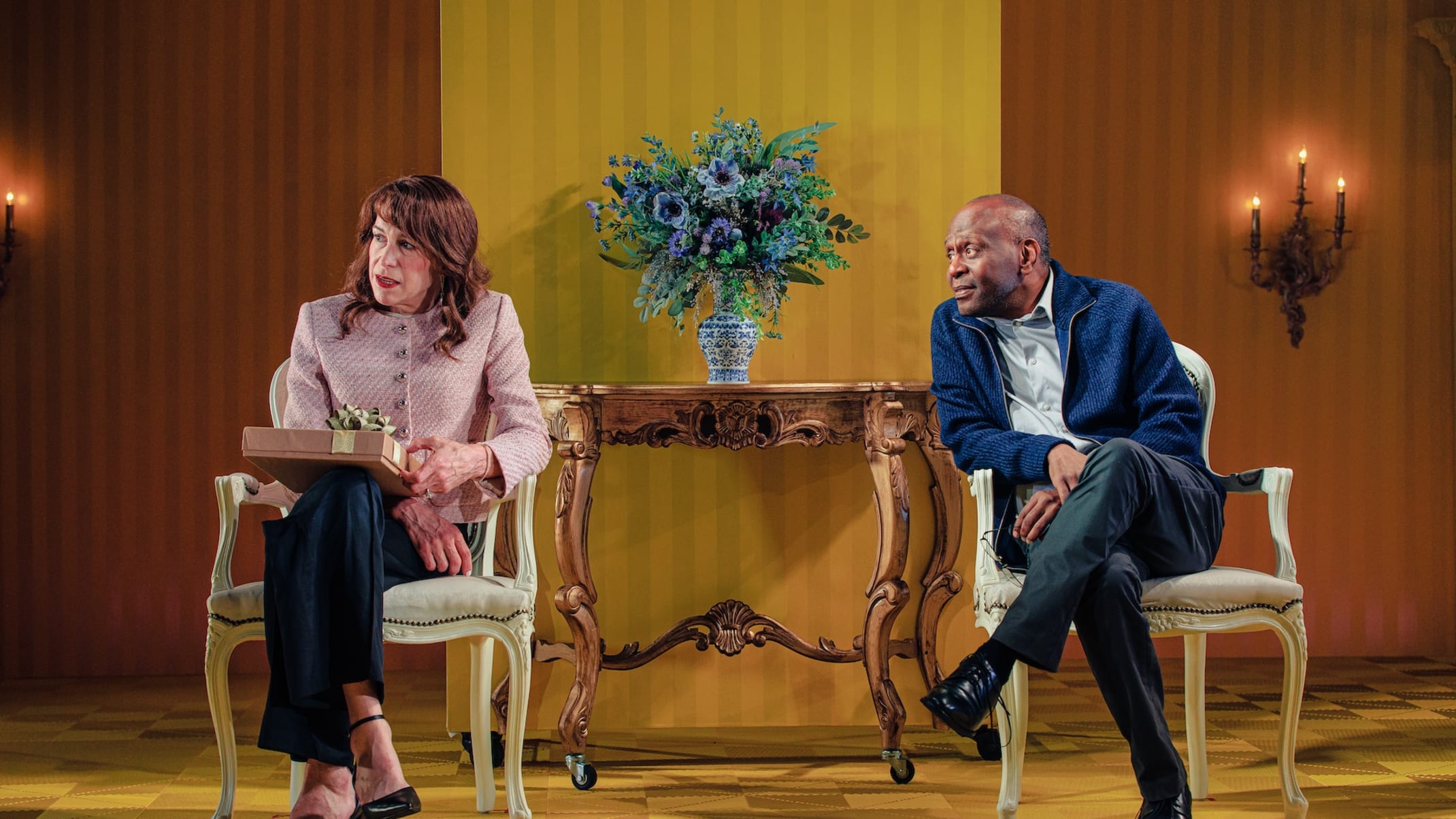 A white woman with brown hair and a bald Black man sit in chairs with their legs crossed.