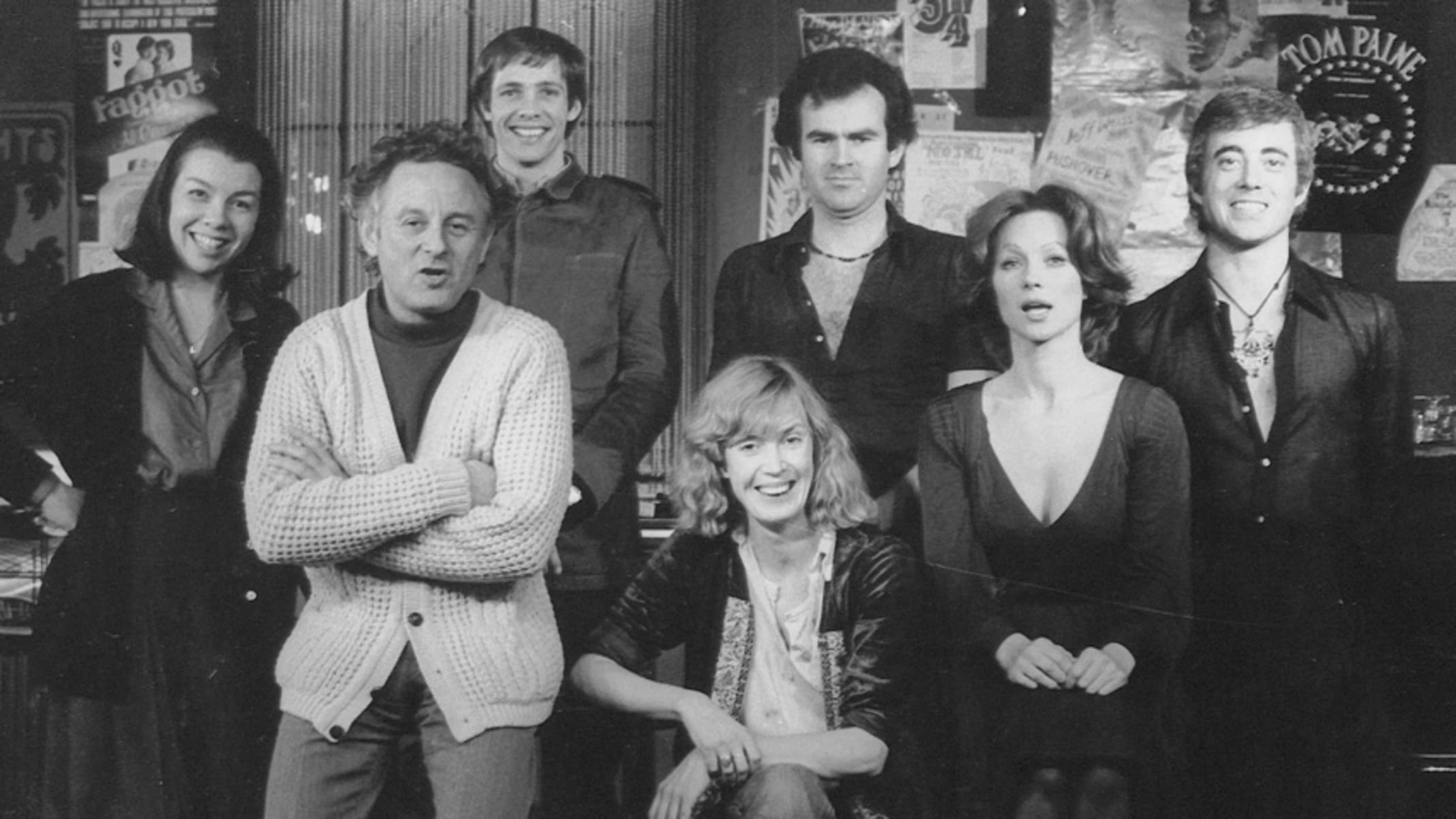 Black and white photo of the cast of Kennedy's Children. The actors stand in front of a wall with posters and other memorabilia.