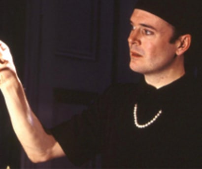 Actor Jefferson Mays in I Am My Own Wife. Mays, a white man, wears a black collared shirt, black head scarf, and a pearl necklace. He looks at his glasses in his outstretched hand.
