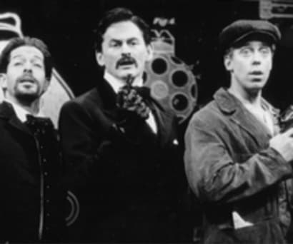 Black and white photo of actors Jonathan Hadary, Victor Garber, and Terrence Mann in Assassins. All three hold guns pointed upwards. Hadary and Garber (both white men) wears suits with leather gloves. Mann (a white man) wears a brown jacket and cap.