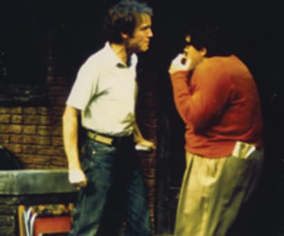 Photos of two actors in Gemini. The man on the right (a white man) wears a white collared shirt and jeans. Standing opposite of him is a white man wearing an orange sweater, tan pants, and glasses; he's cowering in fear.