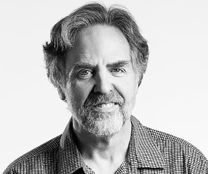 Black and white portrait of Tim Sanford, former Playwrights Horizons Artistic Director, smiling. He wears a plaid collared shirt.