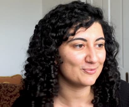 Playwright Sylvia Khoury, a woman of French and Lebanese descent wearing a black top, speaks while seated in her home. A brown pillow and white walls are visible behind her.