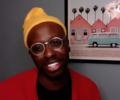 Director and choreographer Raja Feather Kelly, a black man wearing a black top, red sweater, yellow beanie, and glasses, leads a virtual Master Class. A framed photo of a blue van, pink houses, and palm trees hangs on in the wall.
