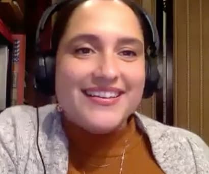 Playwright Jaclyn Backhaus, an Indian-American woman wearing a brown turtleneck, light gray sweater, and large headphones, leads a virtual Master Class.