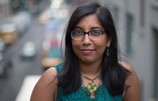 Photo of Natasha Sinha, a South Asian woman in a green top, with a cityscape behind her.