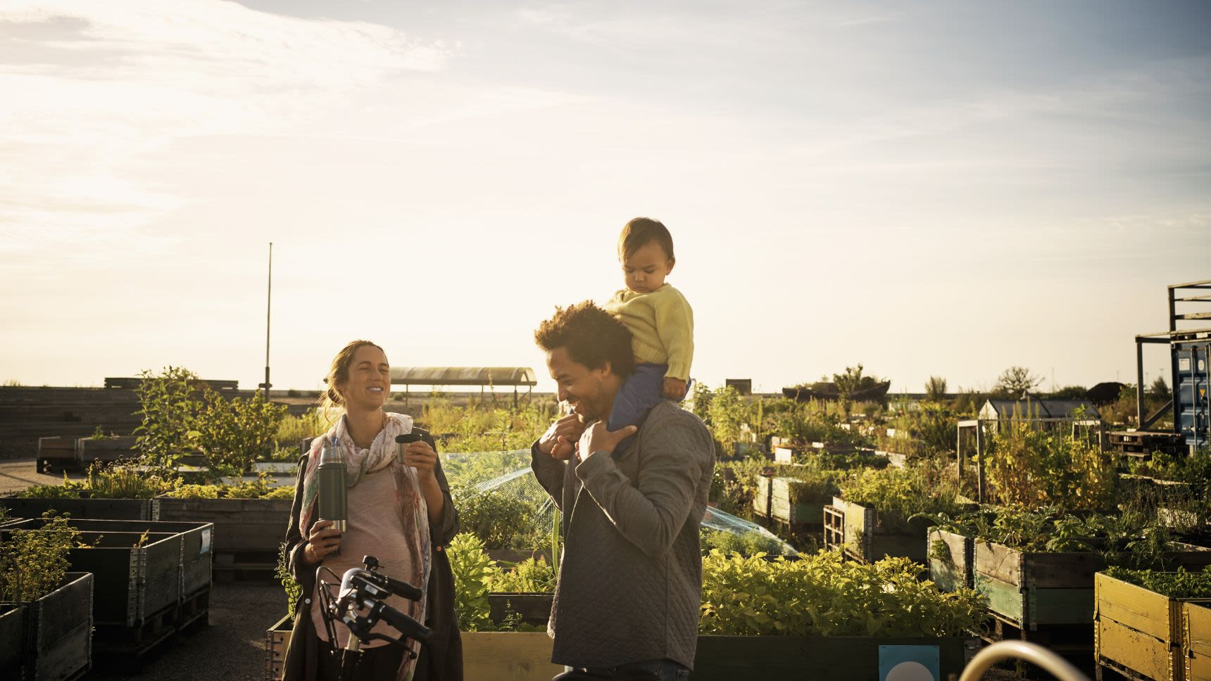 A young family stopping on a bike ride in a community garden where the father holds their young son on his shoulders