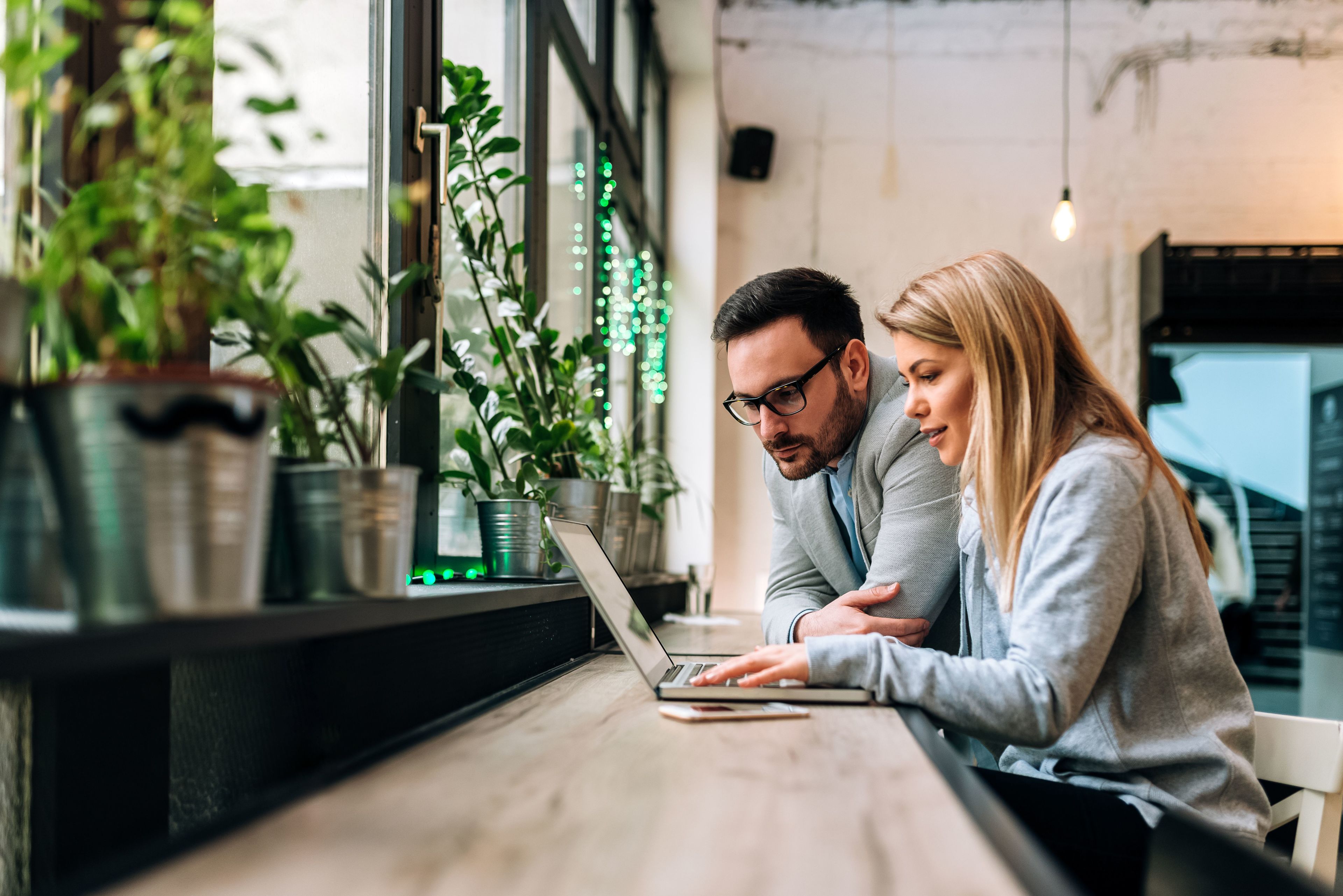 A couple are working together on a laptop in a cafe filled with plants