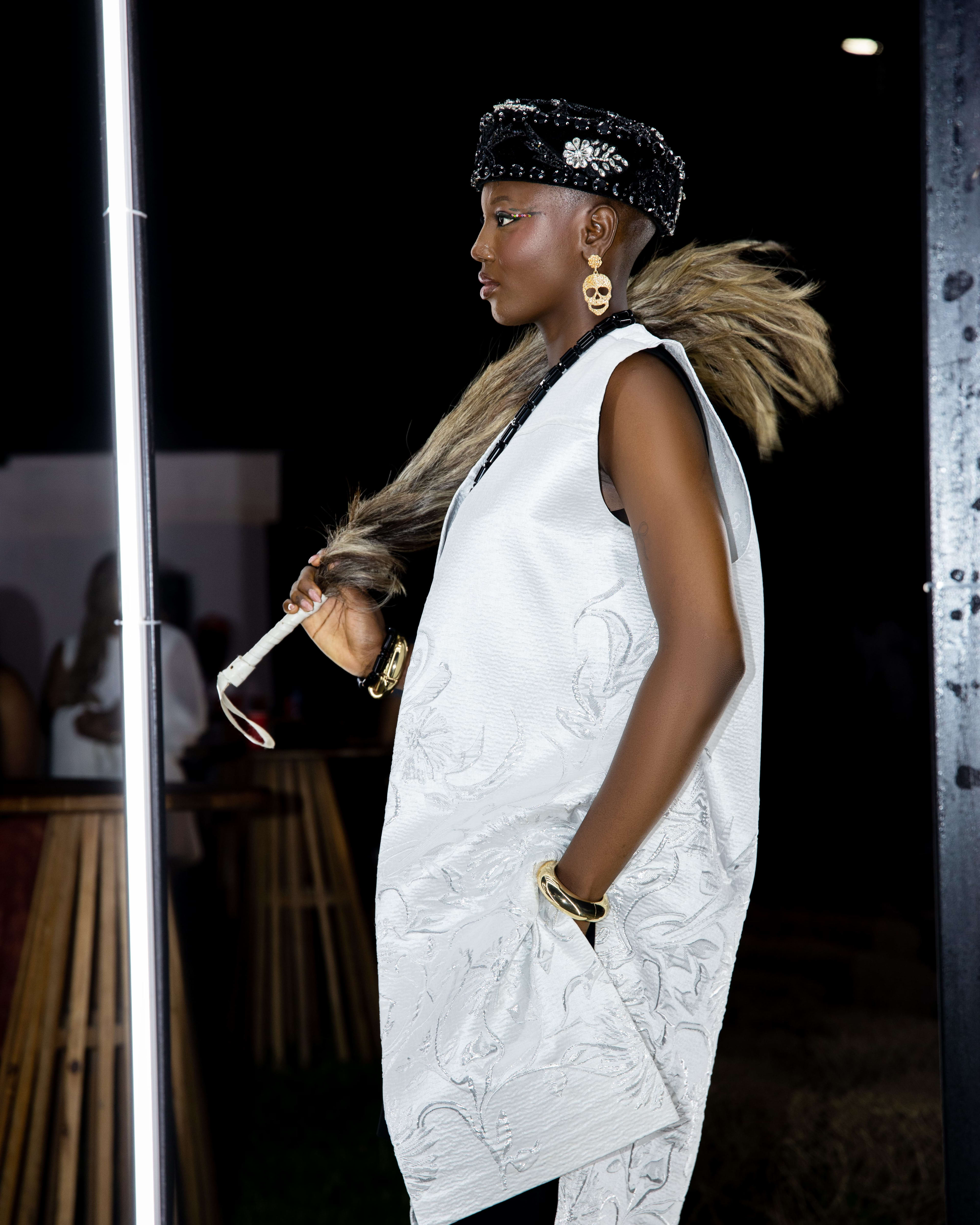A model in a fur-trimmed ivory jacquard vest and beaded cap on the runway, side profile