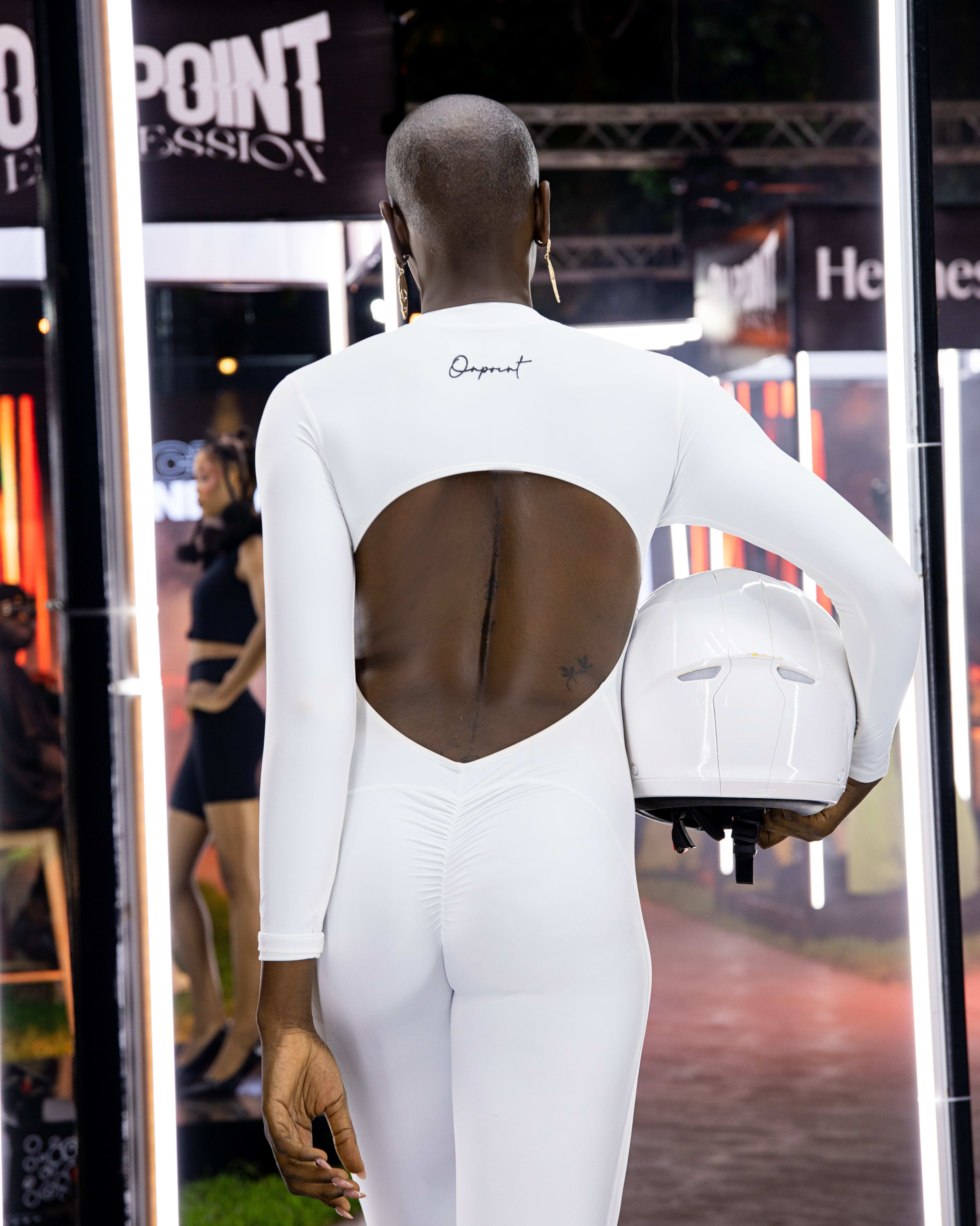 A model in a white cutout bodysuit, back to the camera, carrying a white helmet