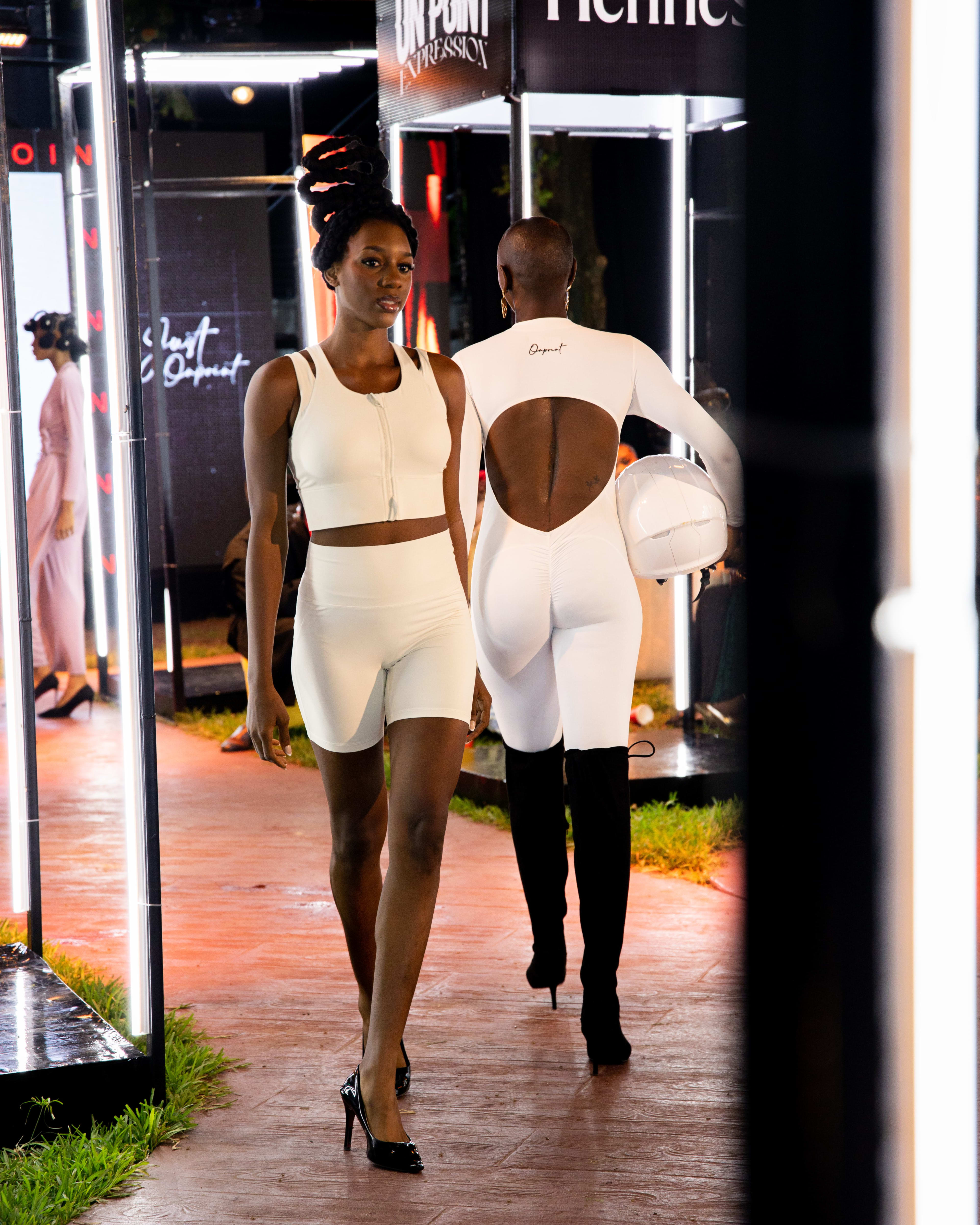 Two models crossing paths on the runway, one in a white cutout bodysuit, one in a matching cream crop set