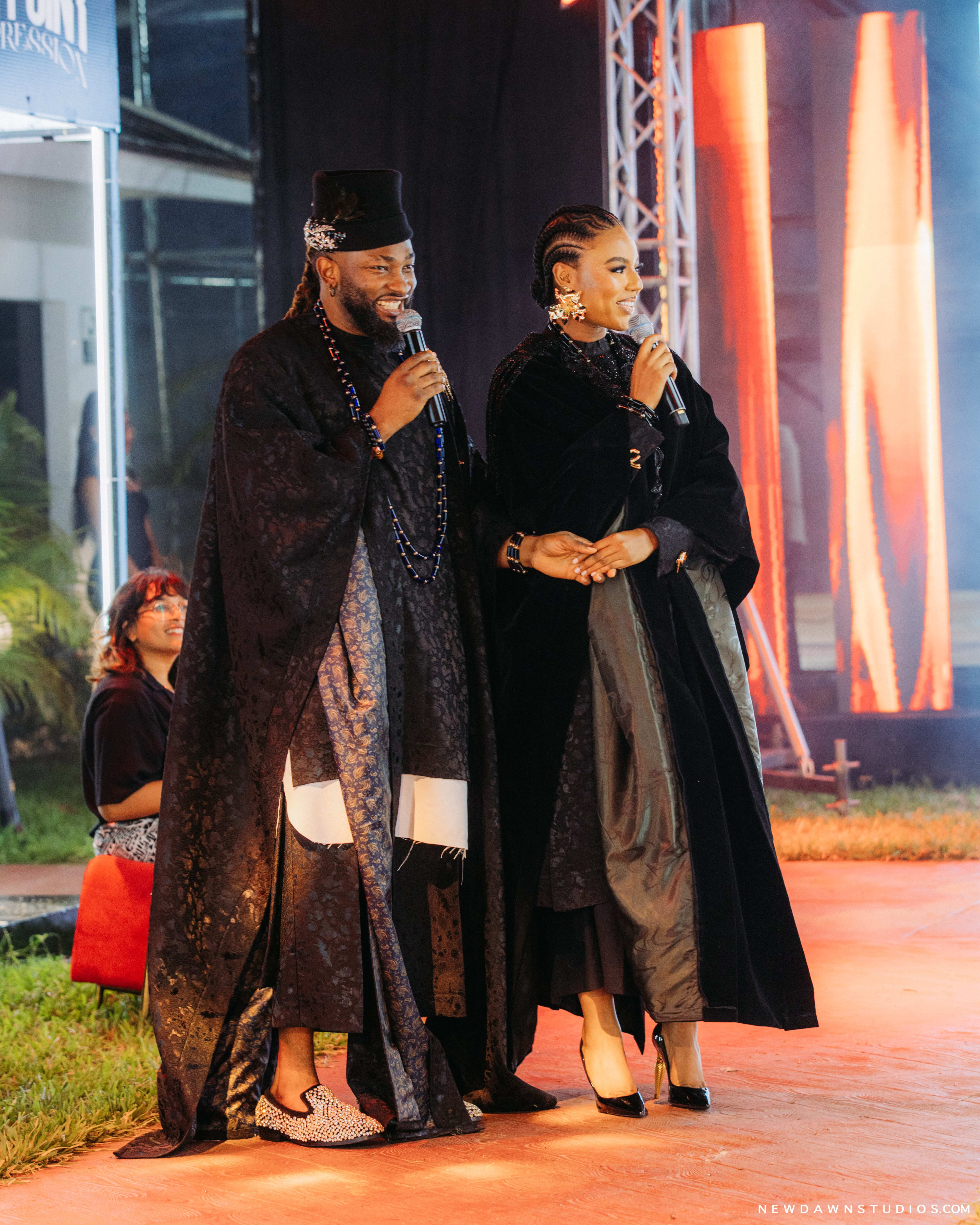 The hosts sharing a warm laugh together, full body, in front of an orange stage curtain