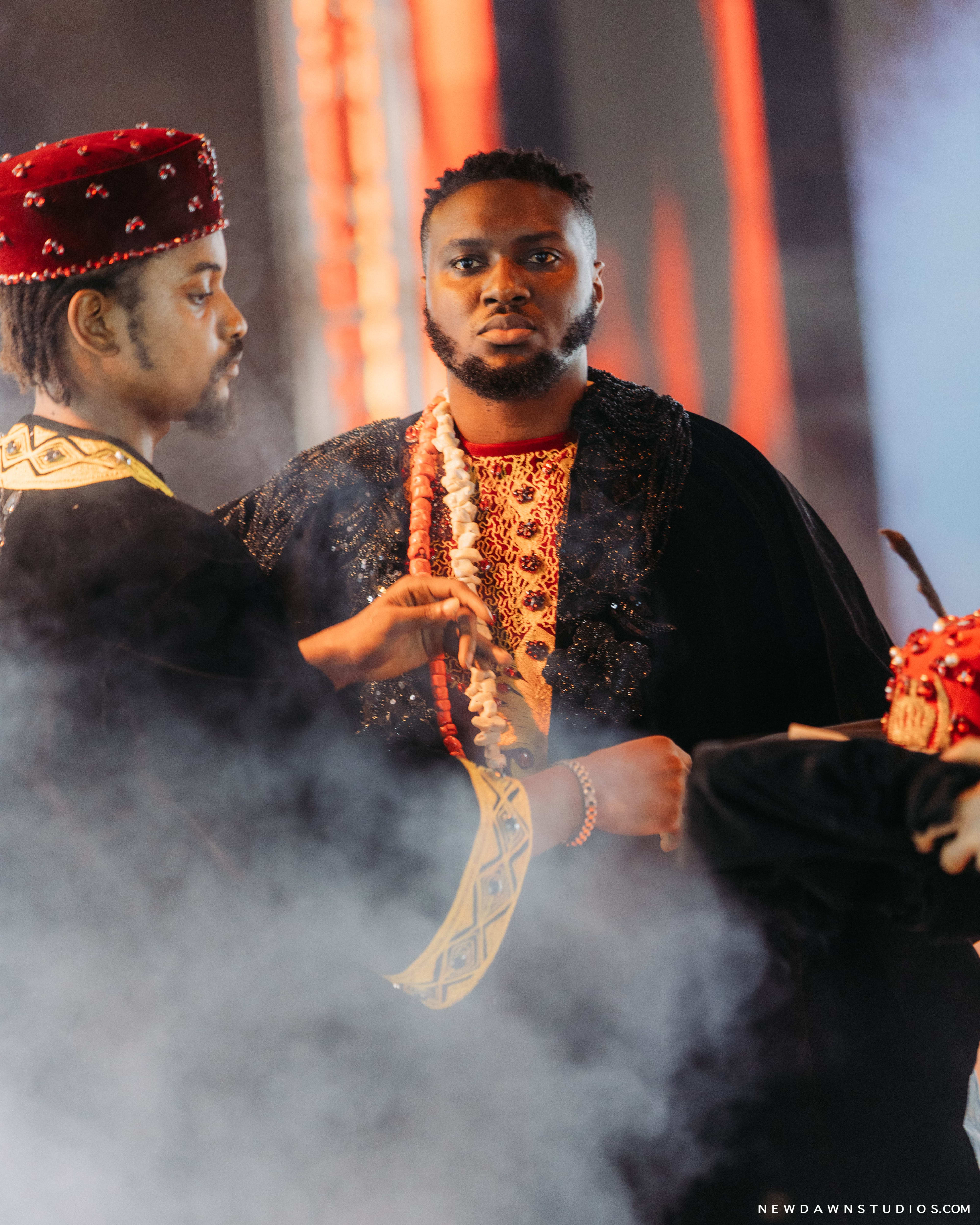 A man in a black velvet cape performing a crowning gesture on another, the pale performer looking on