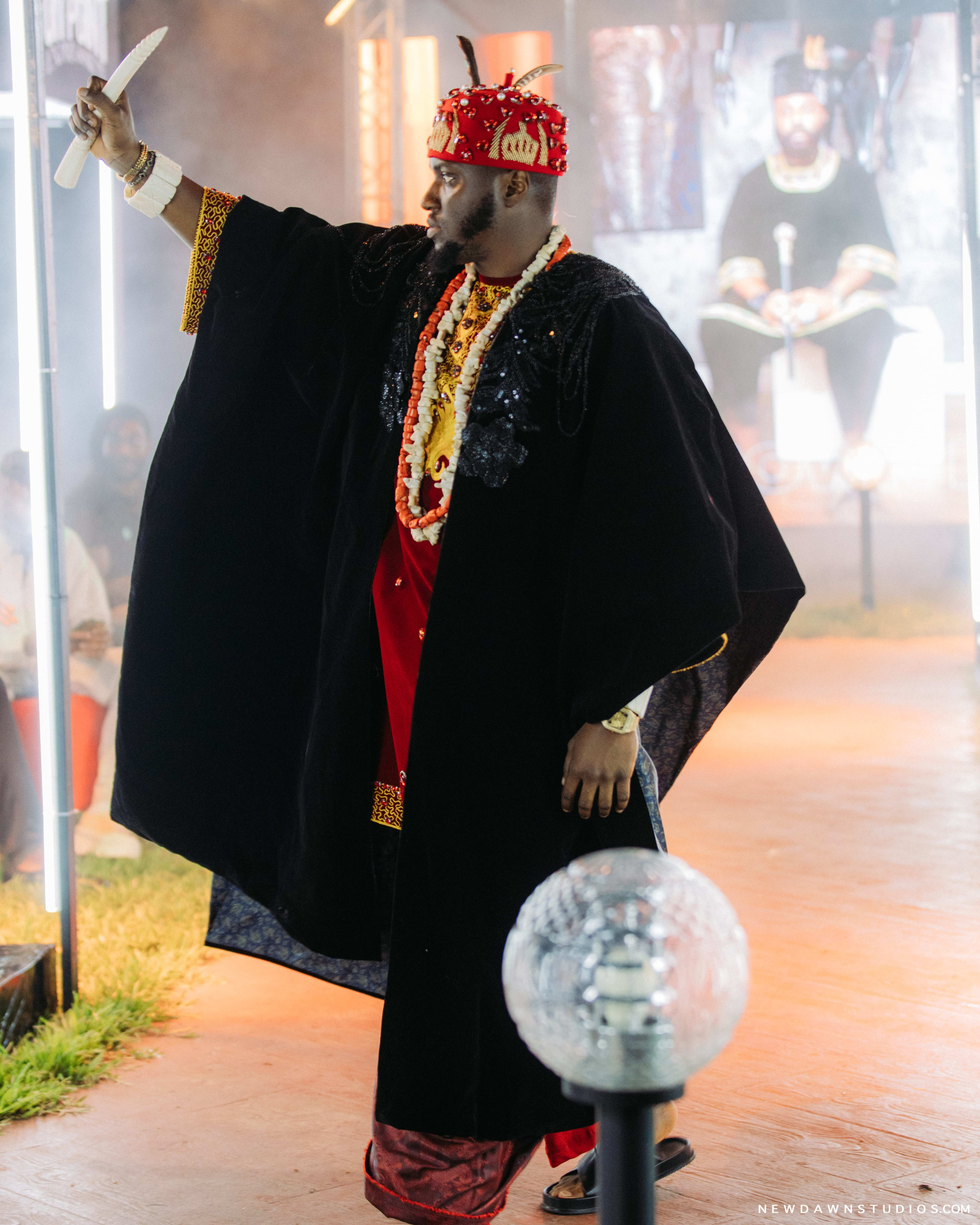 A man in a red cap and black cape raising a curved horn triumphantly overhead amid smoke