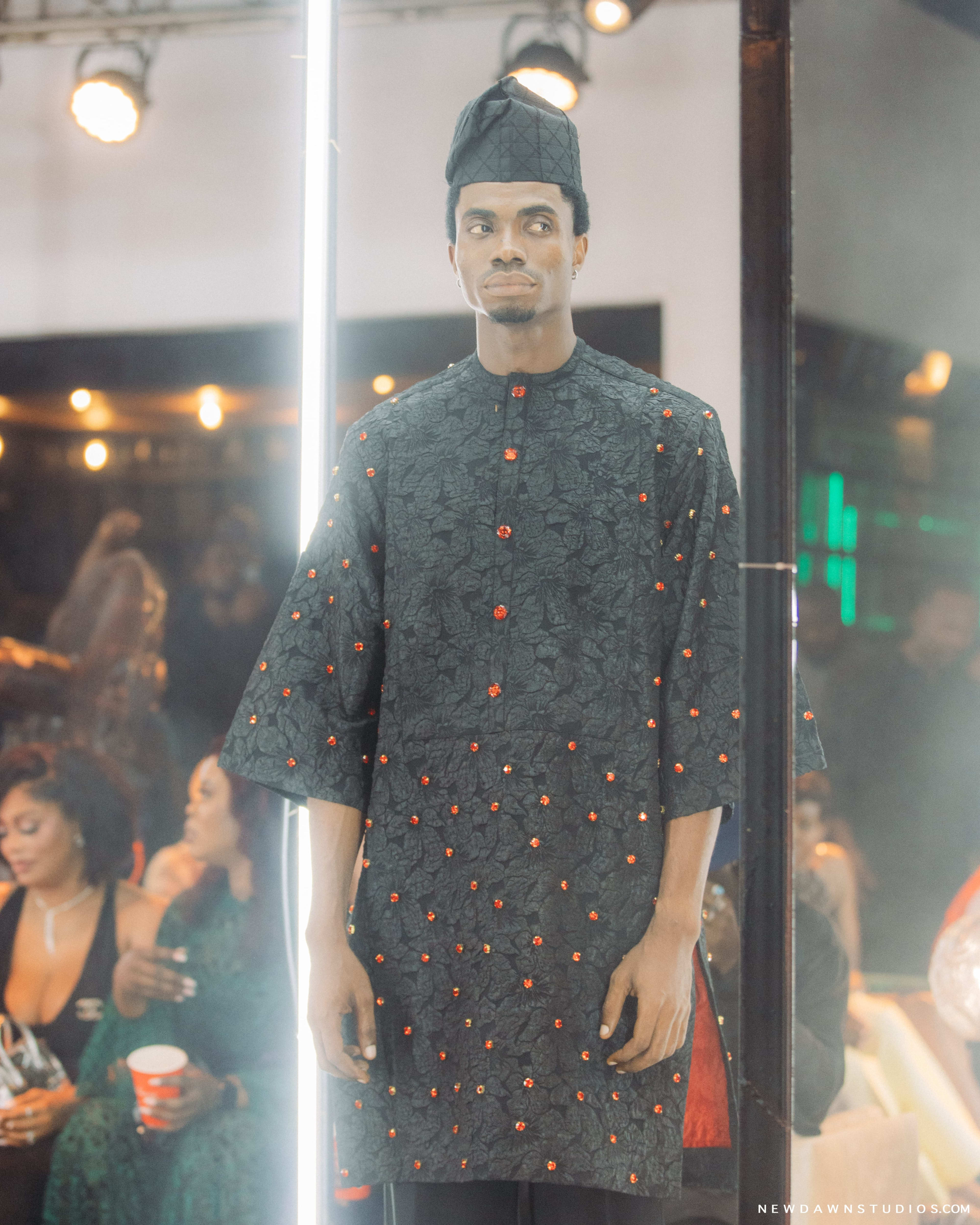 A model in a dark embellished kaftan and cap posed still inside an illuminated mirrored runway box, crowd reflected behind