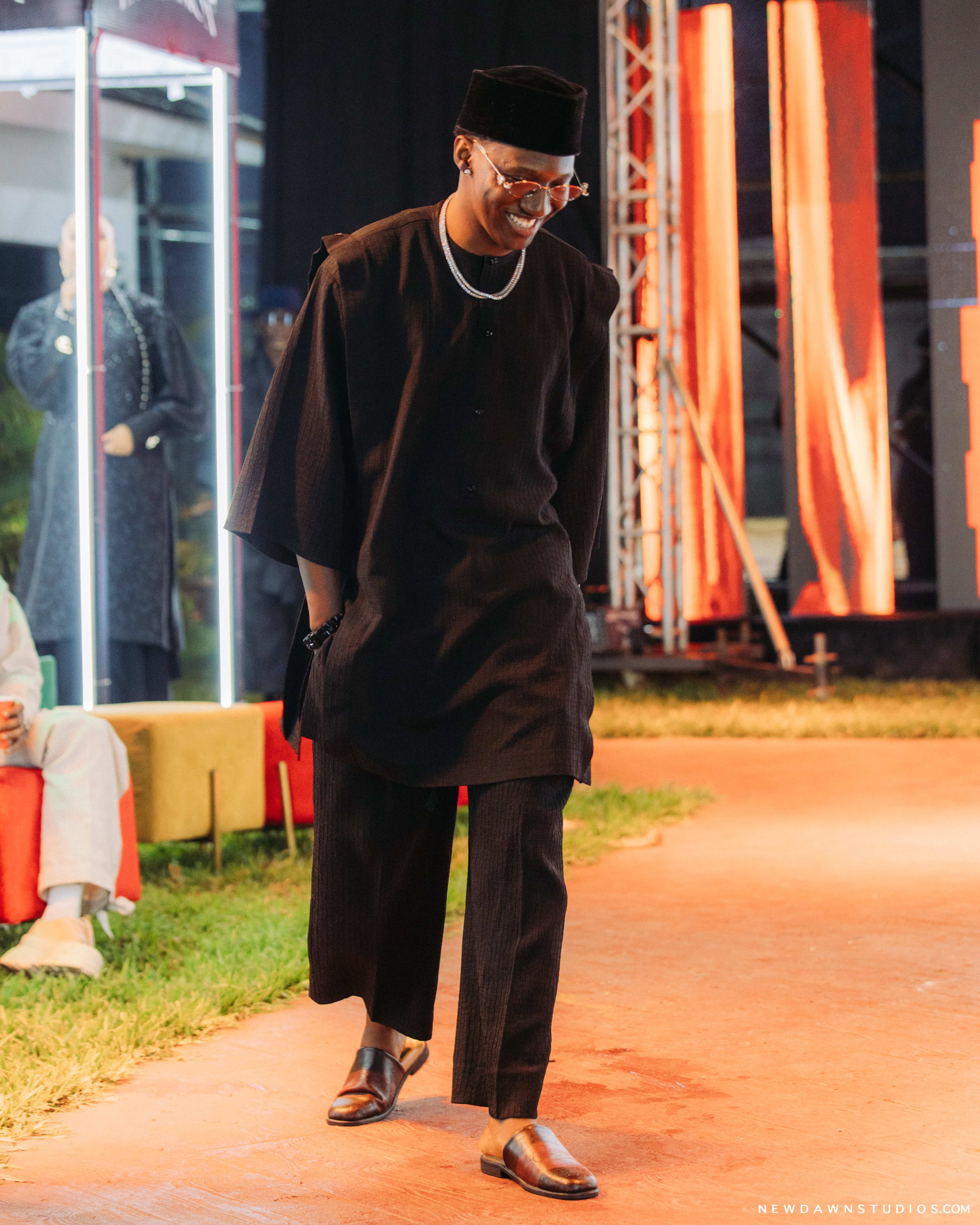 A man in a black kaftan and velvet cap with layered red beads, smiling as he walks the runway