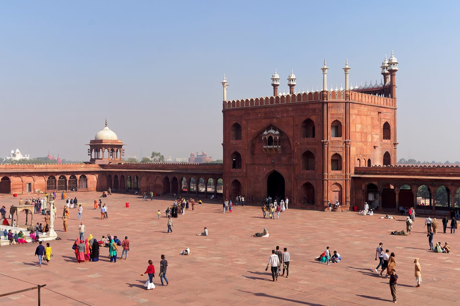 Visitors crossing the wide stone courtyard of Jama Masjid, Delhi, with a red sandstone gate beyond