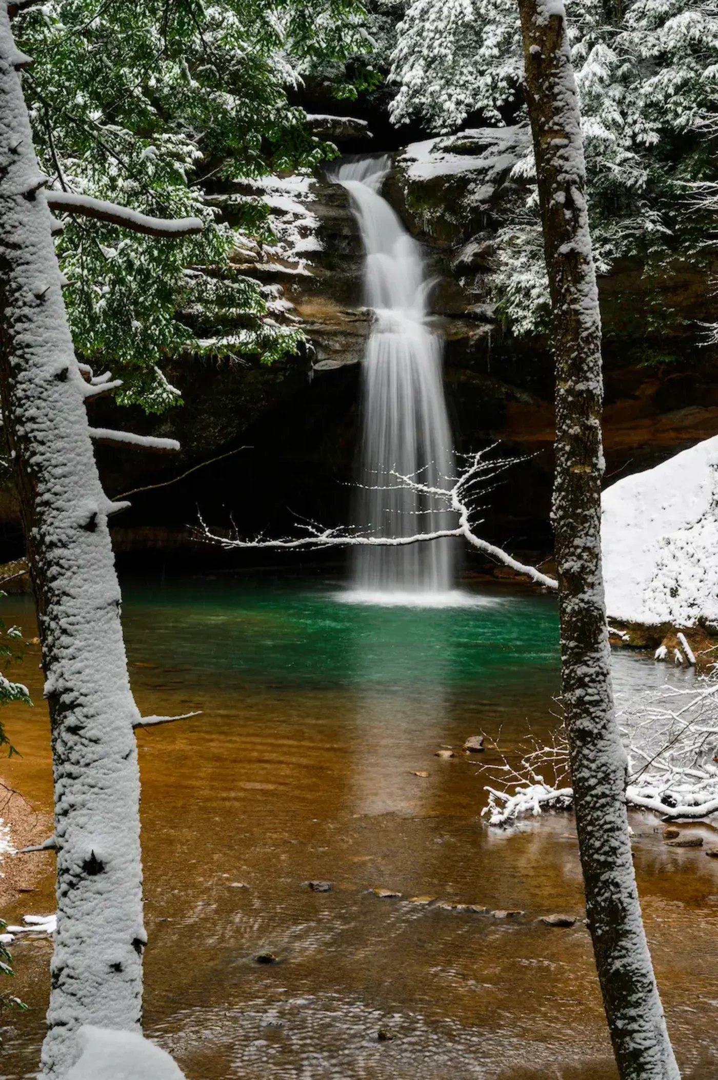 A high-flow snowmelt waterfall into a green pool, framed by snow-dusted trees, Hocking Hills in March