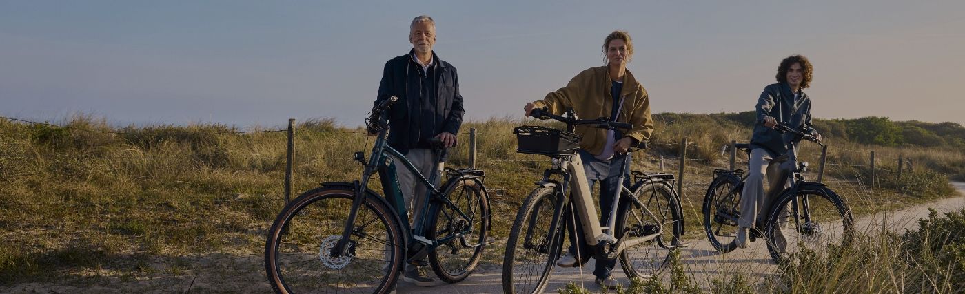Three people walk their bicycles on a sandy path next to grassy dunes at sunset.
