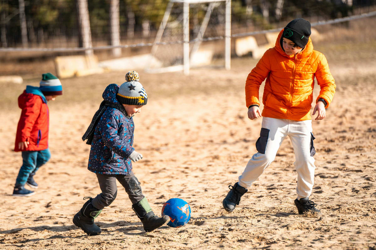 Three children play soccer on a sandy field; one child in a blue coat kicks a blue ball while another in an orange jacket prepares to intercept.