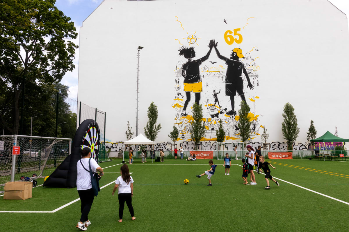 A community sports field with children playing football in the foreground, while adults and other children gather around activity stations. In the background, a large mural on a white building shows two illustrated figures high‑fiving, surrounded by yellow accents and the number 65. Trees, tents, and sports equipment are set up around the field on a sunny day.
