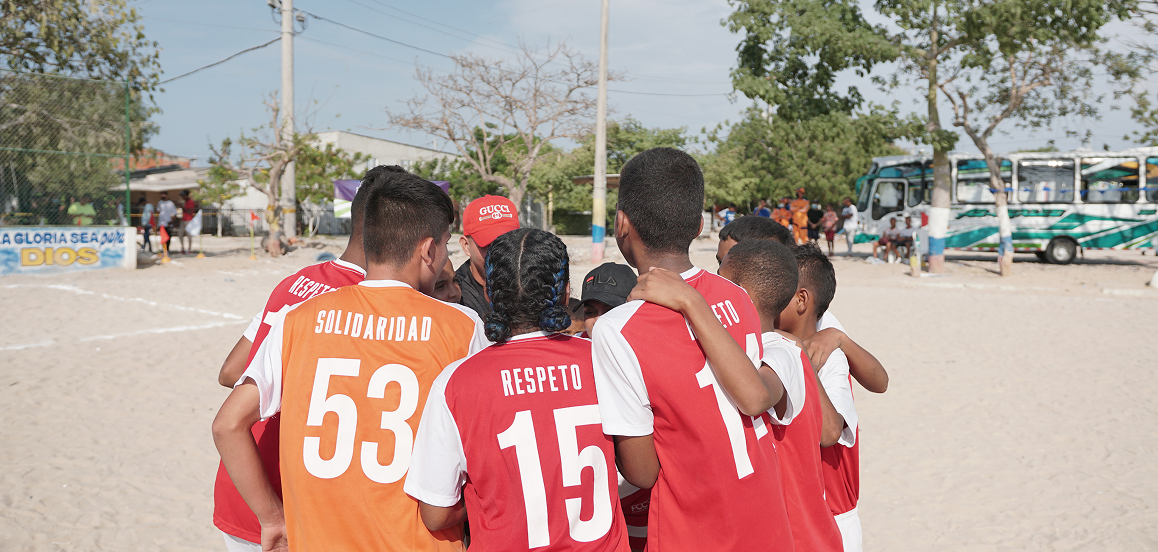 A group of children some in red and some in orange jerseys huddle together on a sandy field. Buses and trees are visible in the background.
