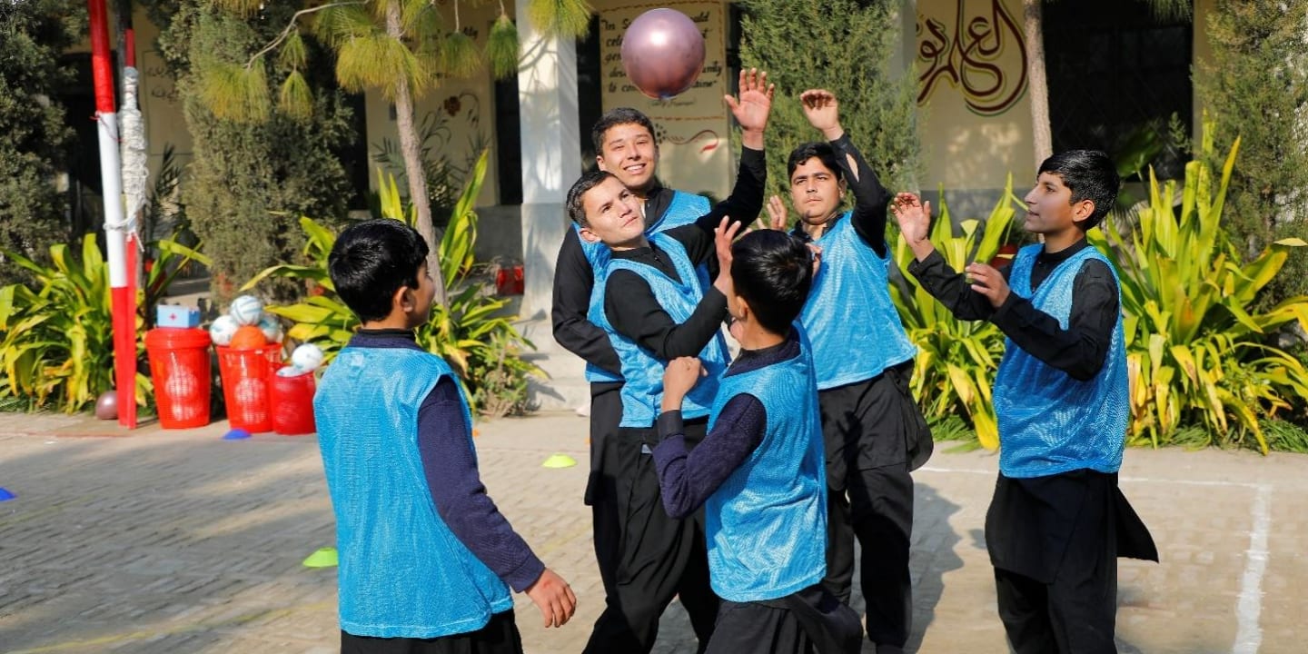 A group of boys wearing black outfits and blue vests play with a ball outdoors on a sunny day.
