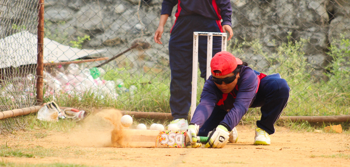 A cricket player in a red cap and sunglasses dives to catch the ball, while another player in matching gear watches. Dust rises from the ground, showing motion.