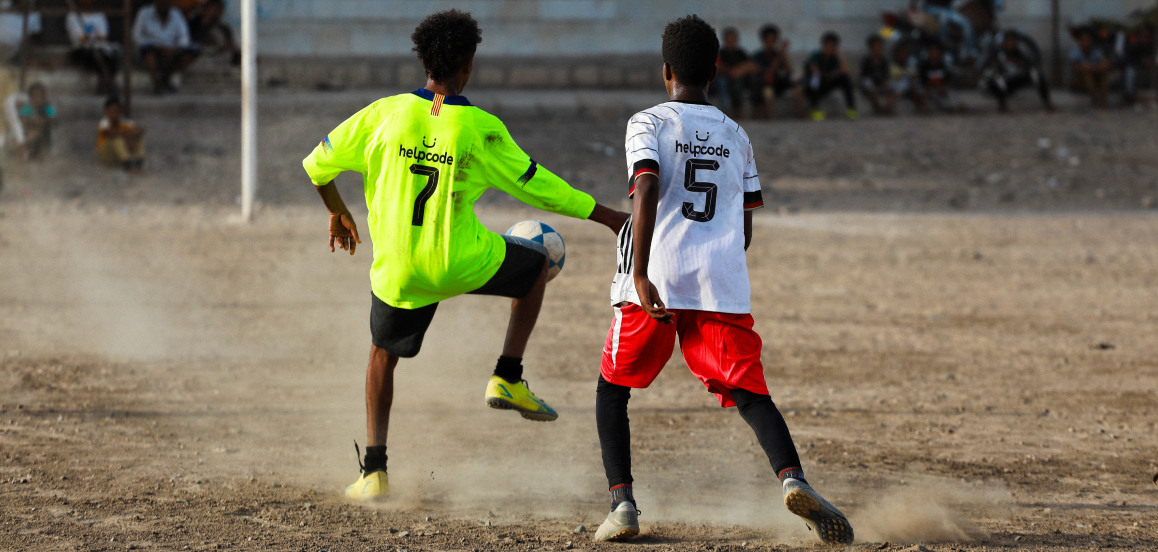 Two young soccer players on a dusty field, one in a neon jersey stopping the ball; others watch from the building in the background.