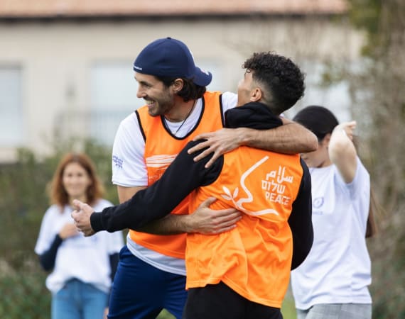 Two young men wearing orange vests, one with PEACE written in English, Hebrew, and Arabic, happily hug during an outdoor activity, while others in white shirts smile in the background.