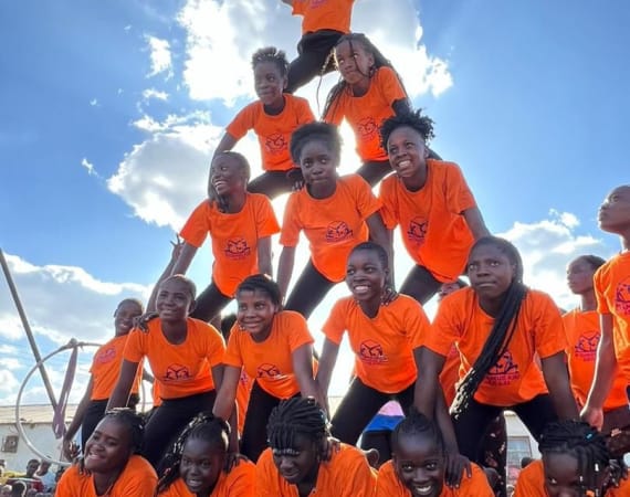A group of children in bright orange T-shirts form a human pyramid under a sunny blue sky.