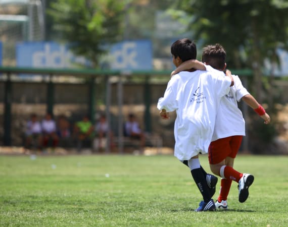 Two young boys wearing sports clothes run together on a grassy field with their arms around each other’s shoulders, suggesting friendship and teamwork, while people sit in the background.