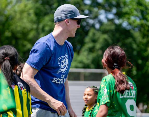 A coach in a blue shirt and cap encourages young soccer players in vibrant yellow and green jerseys on a sunny day.