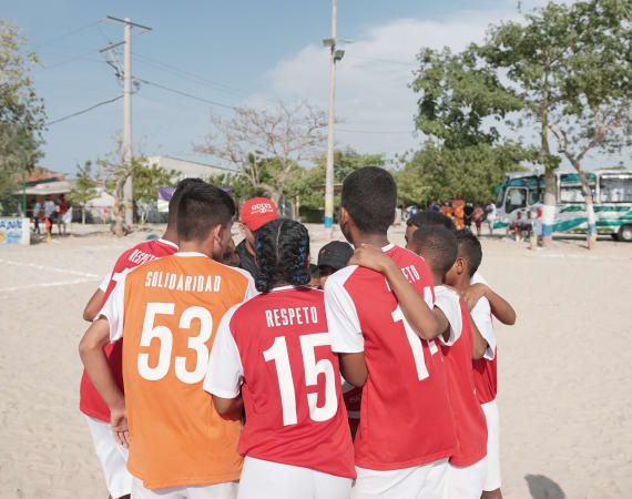 A group of children some in red and some in orange jerseys huddle together on a sandy field. Buses and trees are visible in the background.