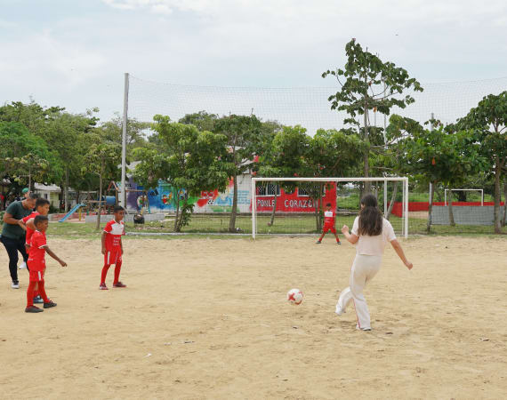 A woman in white kicks a soccer ball towards a goal on a sandy field. Several children in red uniforms watch, with trees and murals in the background.