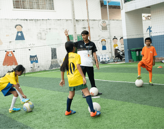 Two girls in yellow jerseys and a boy in orange play soccer on a green field with a coach guiding them.