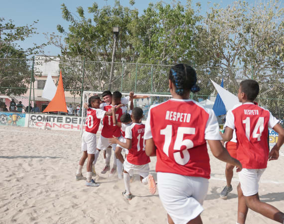 Young soccer players in red and white jerseys celebrate on a sandy field. Banners and trees are visible in the background.