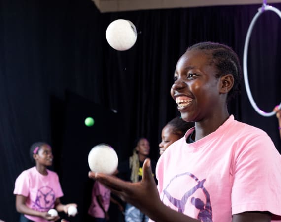 A young person in a pink shirt smiles joyfully while juggling white balls. Others in the background also wear pink shirts.
