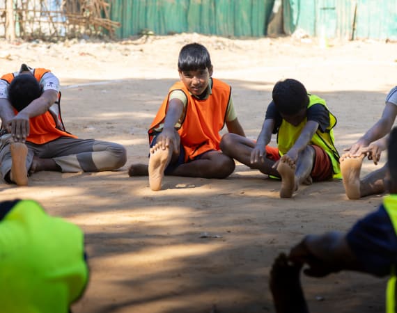 A group of boys wearing bright sports pinnies stretch on a sandy outdoor field, each reaching towards their toes while sitting with legs extended. Sunlight casts shadows on the ground.
