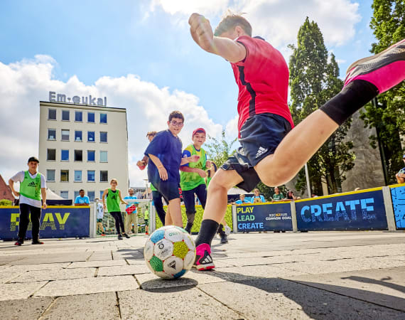 A boy in a red shirt is about to kick a soccer ball on an outdoor court, while other kids in green and purple shirts watch and play under a partly cloudy sky. A building with Em-eukal signage is in the background.