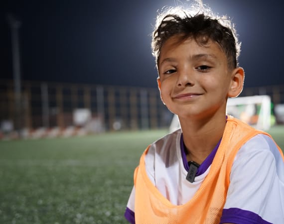 Young boy in an orange sports bib smiles warmly on a dimly lit soccer. The field is empty in the background.