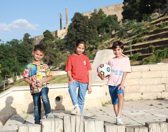Three smiling children pose outdoors on stone steps with a soccer ball. They are surrounded by green trees and an ancient structure in the background.